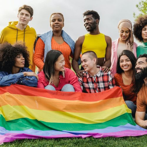Happy diverse friends holding LGBT rainbow flag having fun together outdoors - Focus on bald girl
